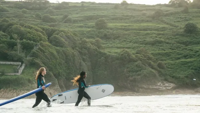 Zwei Anfängerinnen gehen in einem Surfcamp in Spanien mit ihren Surfboards ins Wasser