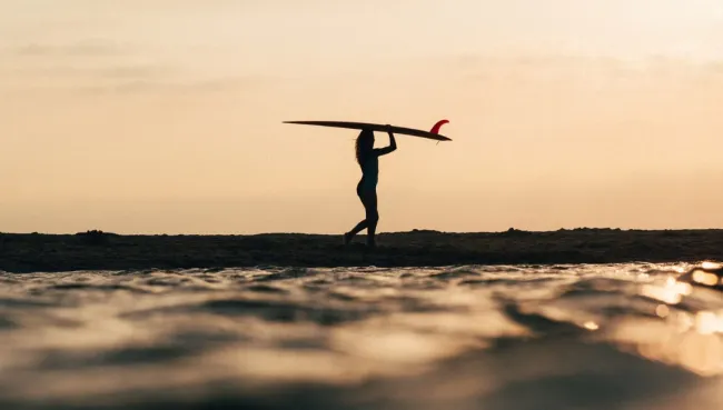 Frau läuft mit ihrem Surfboard bei Sonnenaufgang den Strand entlang