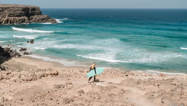 Frau läuft mit ihrem Surfboard an den Strand mit Blick auf die Wellen auf Fuerteventura
