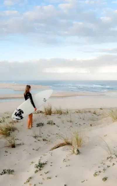 Frau steht mit ihrem Surfboard auf den Dünen in Moliets und schaut auf den Ozean und die Wellen