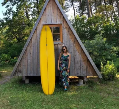 Frau mit ihrem Surfboard vor nachhaltiger Holzhütte im Surfcamp in Frankreich