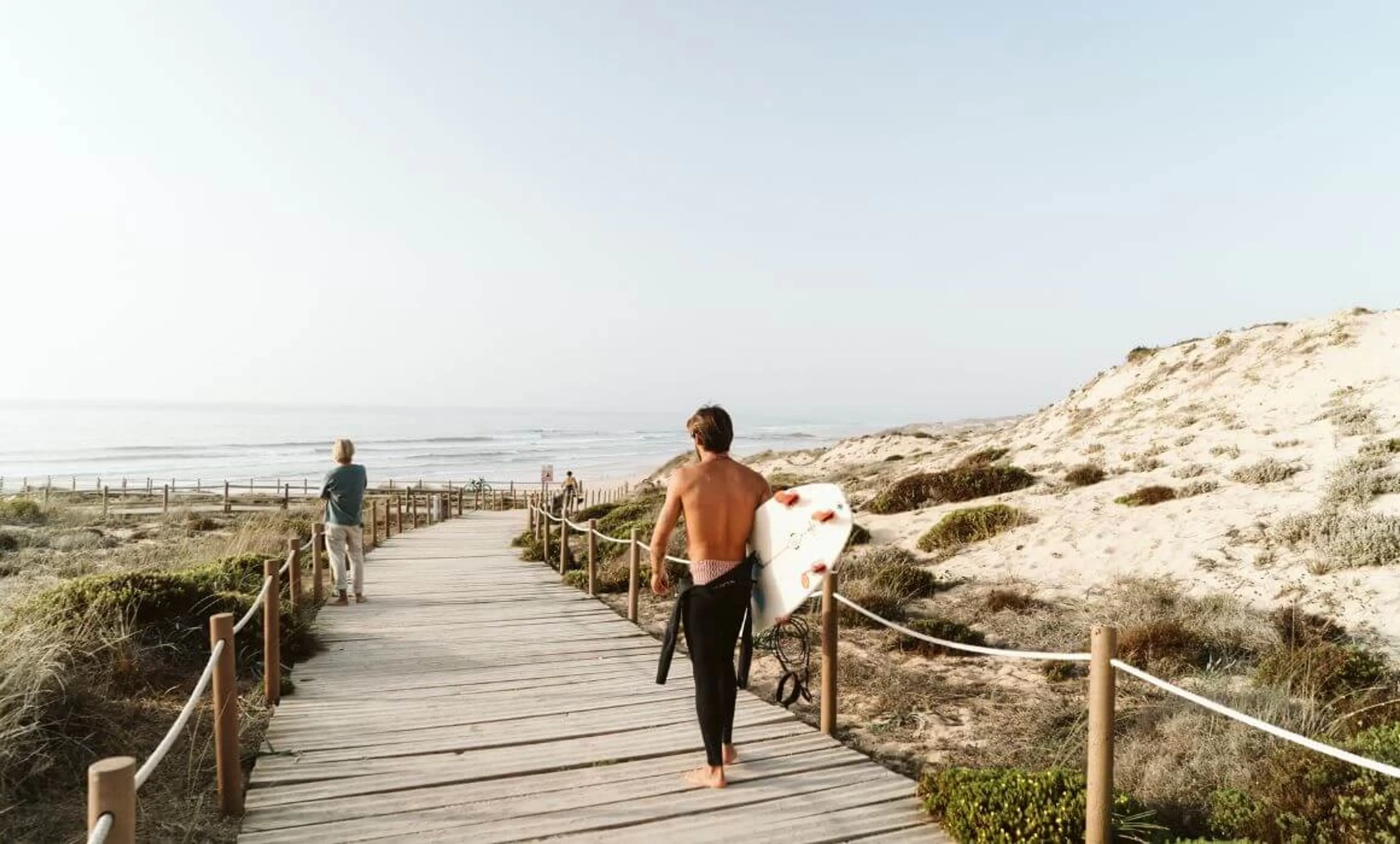 Surfer läuft im Neoprenanzug mit seinem Surfboard den Steg entlang zum Meer