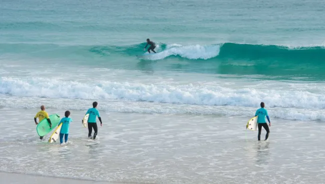 Anfänger surft Welle auf Fuerteventura, während eine Surfcamp-Gruppe ins Meer läuft