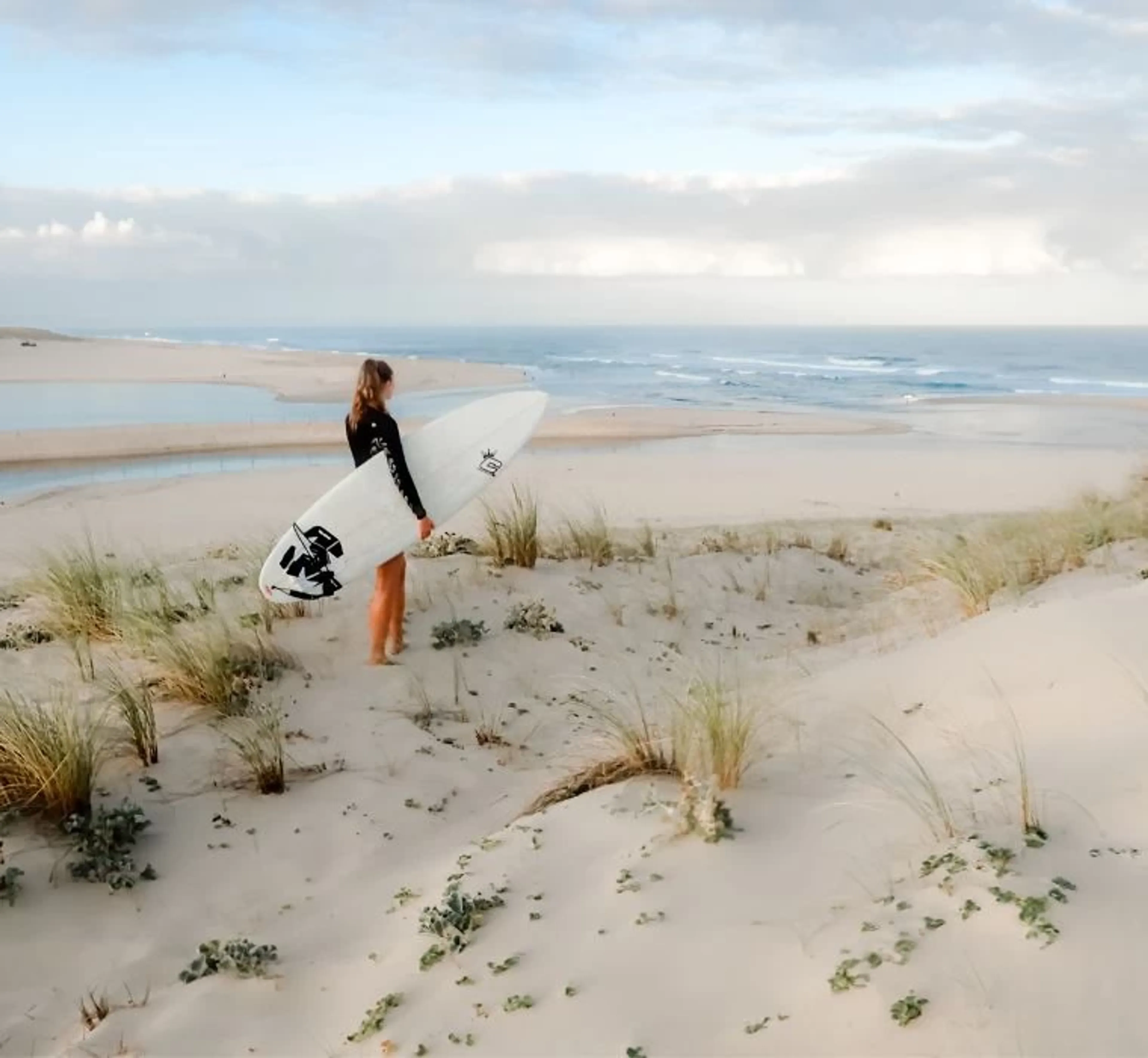Frau mit Surfboard steht auf den Dünen in Frankreich und blickt auf das Meer