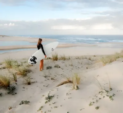 Frau mit Surfboard steht auf den Dünen in Frankreich und blickt auf das Meer