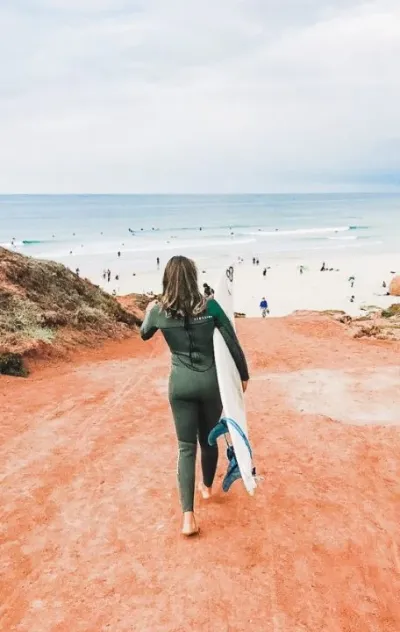 Frau läuft im Neoprenanzug mit ihrem Surfboard zum Strand in Peniche