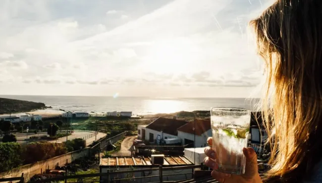 Frau trinkt im Surfcamp in Ericeira einen Cocktail bei Sonnenuntergang mit Blick auf den Strand