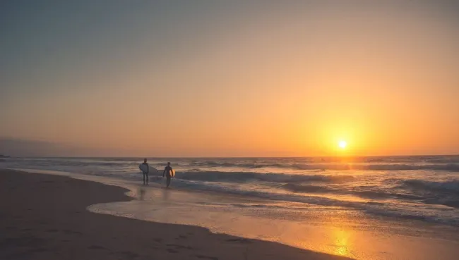 Zwei Surfer laufen mit ihren Surfboards bei Sonnenuntergang den Strand auf Fuerteventura entlang