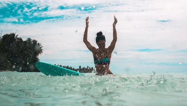 Frau sitzt vor tropischer Kulisse auf Bali lächelnd auf ihrem Surfboard und streckt die Arme hoch
